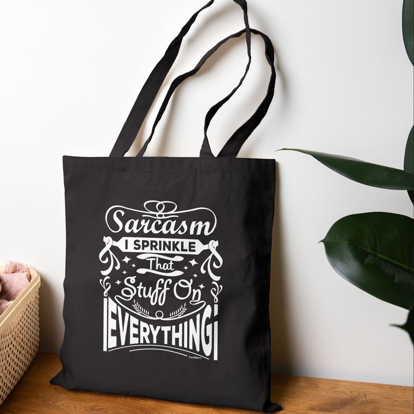 Black tote bag with white text on a wooden surface with a plant in the background