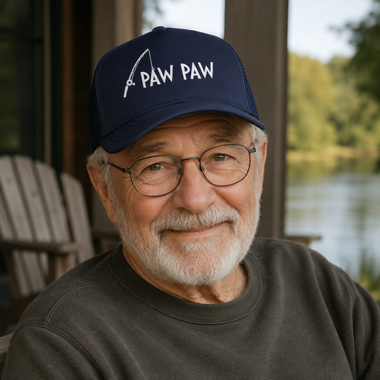 Man wearing a navy blue cap with 'PAW PAW' text and a fishing rod graphic, sitting outdoors by a body of water.